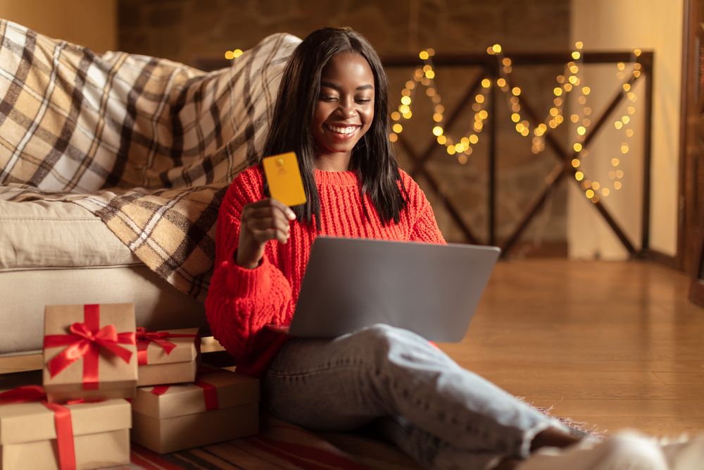 A woman beside gifts, holding a laptop and credit card