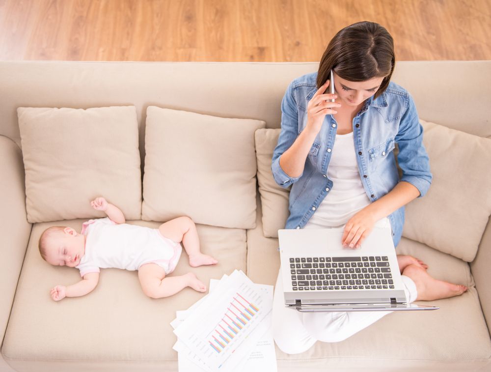 A woman, on a laptop and phone, with her sleeping baby